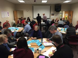 Many adults sit in groups around a room.